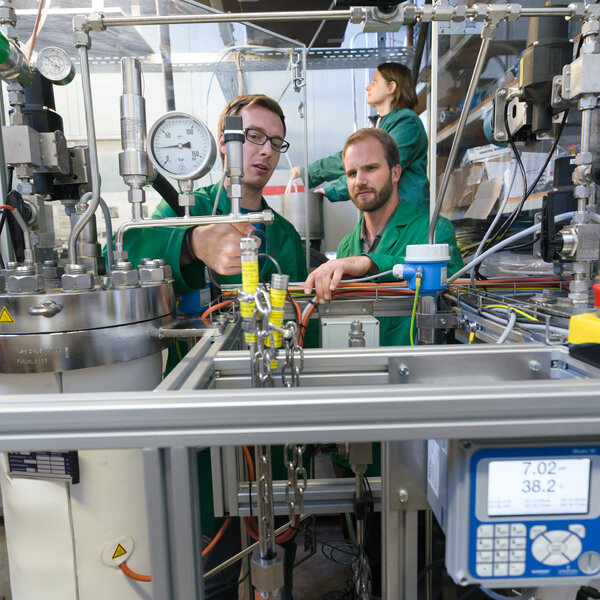 Foto: Drei Personen in grünen Kitteln arbeiten in einem Labor an einer komplexen technischen Anlage mit Druckanzeigen und Schläuchen. Ein Display zeigt "7.02" und "38.2°C". Die Umgebung ist industriell mit zahlreichen Messgeräten und Kabeln.