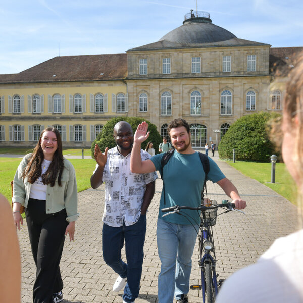 Drei Studierende vor dem Schloss Mittelbau der Universität Hohenheim, einer winkt in die Kamera.