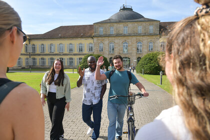 Drei Studierende vor dem Schloss Mittelbau der Universität Hohenheim, einer winkt in die Kamera.