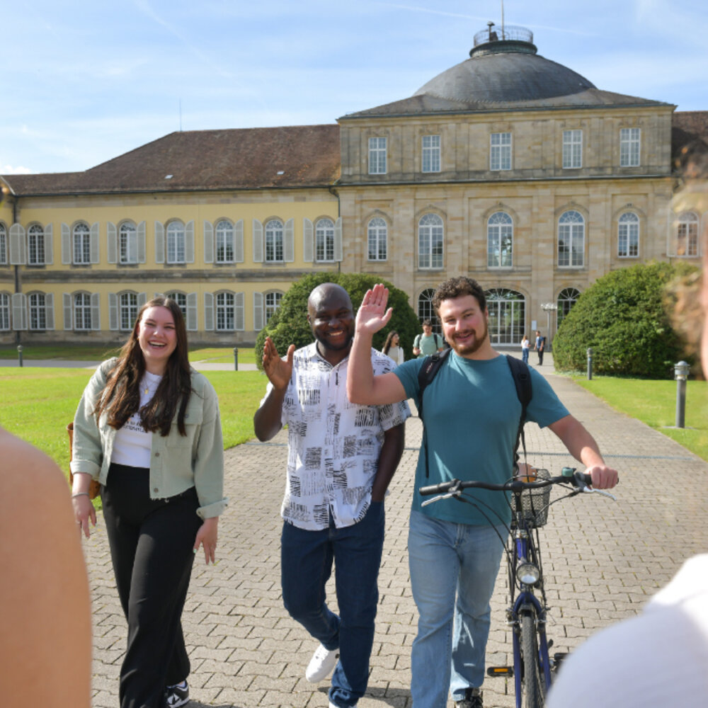 Drei Studierende vor dem Schloss Mittelbau der Universität Hohenheim, einer winkt in die Kamera.