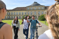 Drei Studierende vor dem Schloss Mittelbau der Universität Hohenheim, einer winkt in die Kamera.