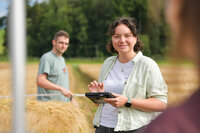 Studentin steht auf Getreidefeld mit Tablet. Im Hintergrund steht ein Student zwischen den Ähren.