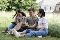 Foto: Drei Personen sitzen auf einer Wiese und nutzen Tablets. Eine Person schreibt mit einem Stift auf einem Tablet, während die anderen beiden ein weiteres Tablet betrachten. Im Hintergrund sind Bäume und ein Gebäude mit Schieferdach sichtbar.