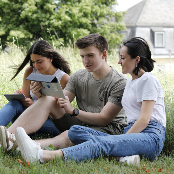 Foto: Drei Personen sitzen auf einer Wiese und nutzen Tablets. Eine Person schreibt mit einem Stift auf einem Tablet, während die anderen beiden ein weiteres Tablet betrachten. Im Hintergrund sind Bäume und ein Gebäude mit Schieferdach sichtbar.