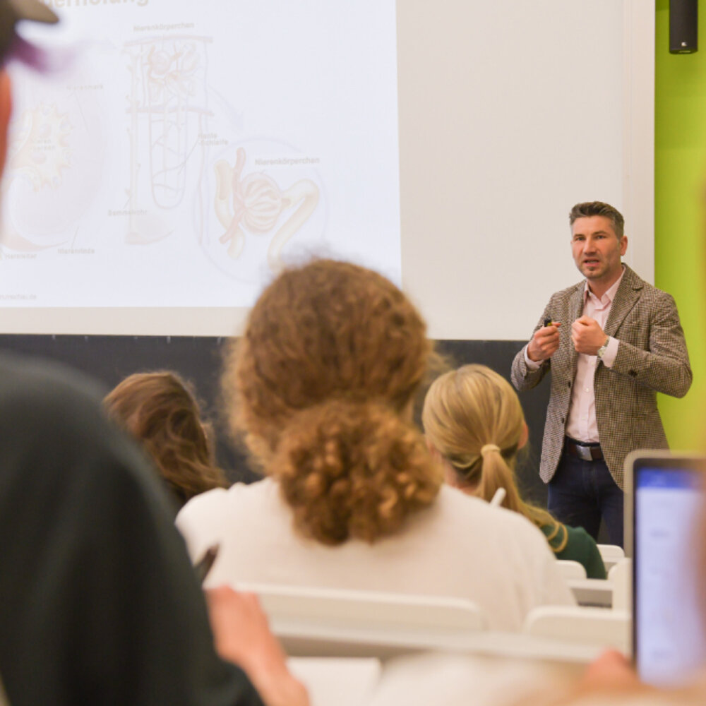 Dozent spricht vor Studierenden in einem Hörsaal mit Präsentation im Hintergrund.