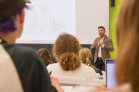 Dozent spricht vor Studierenden in einem Hörsaal mit Präsentation im Hintergrund.