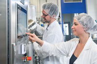 Foto: Zwei Personen in weißen Laborkitteln und Haarnetzen bedienen eine Maschine mit einem Touchscreen in einem Labor. Eine Person zeigt auf den Bildschirm, während die andere an einem Behälter arbeitet. Im Hintergrund sind industrielle Geräte sichtbar.