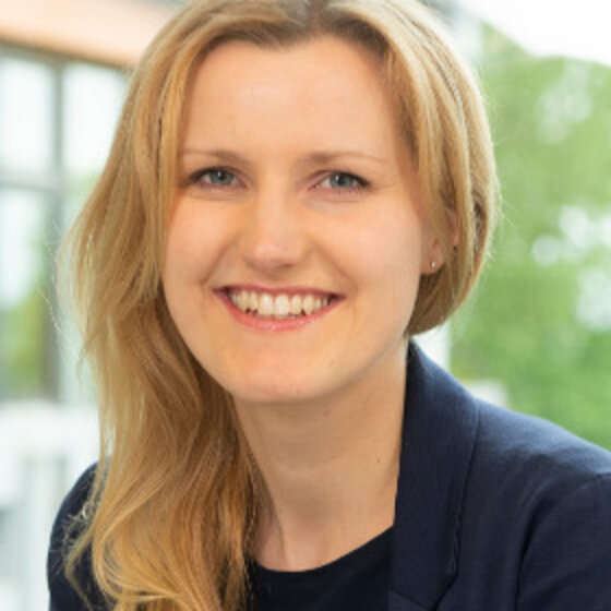 Foto: Eine Person mit langen Haaren steht im Freien vor einem Fenster mit Bäumen im Hintergrund. Der Hintergrund zeigt einen bewölkten Himmel. Die Person trägt ein blaues Oberteil und einen Blazer.