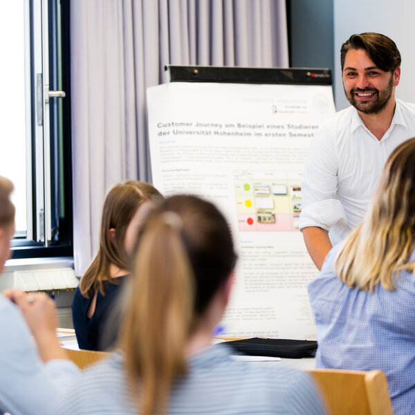Foto: Eine Person steht vor einem Poster mit dem Titel 'Customer Journey am Beispiel eines Studierenden der Universität Hohenheim im ersten Semester'. Mehrere Personen sitzen und hören zu. Der Raum hat ein Fenster mit Vorhängen und es sind weitere Poster im Hintergrund sichtbar.