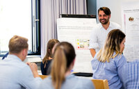 Foto: Eine Person steht vor einem Poster mit dem Titel 'Customer Journey am Beispiel eines Studierenden der Universität Hohenheim im ersten Semester'. Mehrere Personen sitzen und hören zu. Der Raum hat ein Fenster mit Vorhängen und es sind weitere Poster im Hintergrund sichtbar.