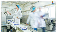 Foto: Eine Person in weißem Kittel und blauer Haube bedient ein Kontrollpanel in einer Fabrikumgebung. Im Vordergrund laufen zwei weitere Personen in ähnlicher Kleidung an einer Produktionslinie vorbei. Maschinen und Förderbänder sind sichtbar.