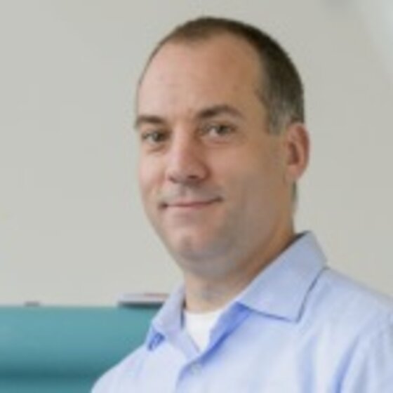 Foto: Eine Person in einem hellblauen Hemd steht vor einer Maschine. Im Hintergrund ist ein gelbes Warnschild mit einem schwarzen Ausrufezeichen sichtbar. Die Umgebung wirkt industriell oder technisch.