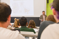 Dozent erklärt anatomischen Zeichnungen Studierenden im Hörsaal, mit Notizblöcken und Laptops.