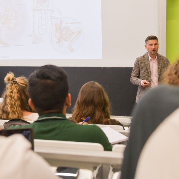 Dozent erklärt anatomischen Zeichnungen Studierenden im Hörsaal, mit Notizblöcken und Laptops.