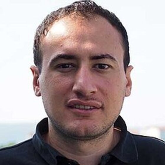 Foto: Eine Person steht im Freien vor einer verschwommenen Landschaft mit Bäumen und einem Gebäude im Hintergrund. Die Person trägt ein dunkles Oberteil. Der Himmel ist klar und hell.