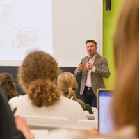 [Übersetzen nach: English] Dozent spricht vor Studierenden in einem Hörsaal mit Präsentation im Hintergrund.