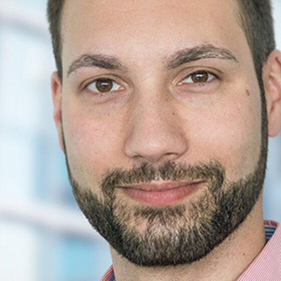 Foto: Eine Person in einem rosa Hemd steht vor einem modernen Gebäude mit Glasfassade. Der Hintergrund zeigt unscharfe Fensterstrukturen.
