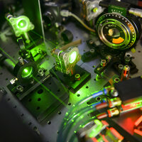 Foto: Eine Nahaufnahme eines optischen Labors mit mehreren Geräten und grünen Laserstrahlen. Verschiedene Linsen und Spiegel sind auf einer perforierten Metallplatte montiert. Ein Gerät zeigt eine Skala mit der Aufschrift "B. Halle". Kabel und Schläuche sind ebenfalls sichtbar.