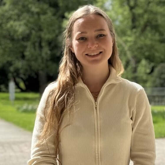 Foto: Eine Person steht auf einem gepflasterten Weg in einem grünen Park mit Bäumen und Rasenflächen im Hintergrund. Die Person trägt eine helle Jacke und hat langes Haar. Der Park wirkt gepflegt und ruhig.