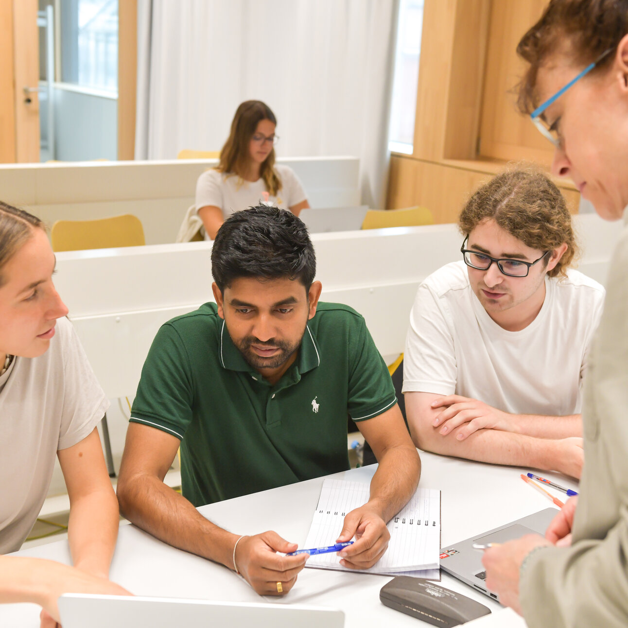 Foto: Mehrere Personen sitzen an einem Tisch in einem Klassenzimmer und arbeiten zusammen. Eine Person steht und scheint Anweisungen zu geben. Auf dem Tisch liegen Notizbücher, Stifte und ein Laptop. Im Hintergrund sind weitere Sitzreihen zu sehen.