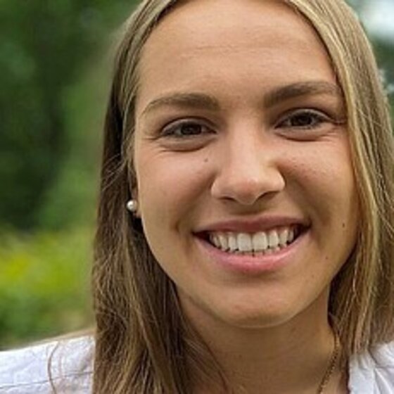 Foto: Eine Person mit langen Haaren steht im Freien vor einer grünen Hecke. Der Hintergrund zeigt Bäume und einen bewölkten Himmel. Die Person trägt ein weißes Oberteil.