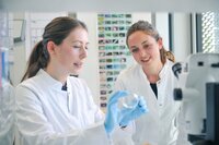 Foto: Zwei Personen in weißen Laborkitteln arbeiten in einem Labor. Eine Person hält eine Petrischale mit blauen Handschuhen, während die andere zuschaut. Im Hintergrund sind ein Mikroskop und eine Wand mit Bildern sichtbar.