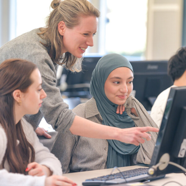 Lehrende zeigt drei Studierenden etwas am Bildschirm in einem Computerraum.