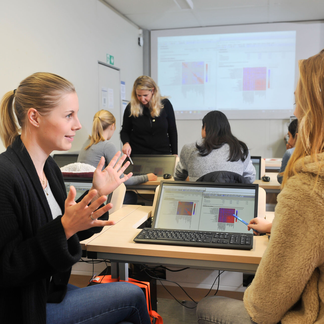 Foto: Mehrere Personen sitzen in einem Schulungsraum und arbeiten an Laptops. Eine Person steht vorne und spricht mit anderen. Auf den Bildschirmen sind Diagramme zu sehen, und im Hintergrund wird eine Präsentation projiziert.