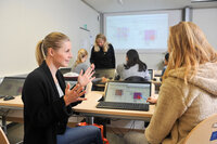 Foto: Mehrere Personen sitzen in einem Schulungsraum und arbeiten an Laptops. Eine Person steht vorne und spricht mit anderen. Auf den Bildschirmen sind Diagramme zu sehen, und im Hintergrund wird eine Präsentation projiziert.