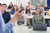 Foto: Eine Gruppe von Personen sitzt in einem Hörsaal und hört einem Vortrag zu. Im Vordergrund ist eine Person mit einem Laptop zu sehen. Der Raum ist mit Tischen und Stühlen ausgestattet.