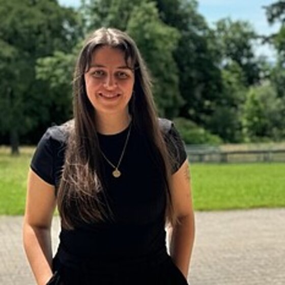 Foto: Eine Person steht auf einem gepflasterten Weg in einem Park, umgeben von grünen Bäumen und Rasenflächen. Im Hintergrund ist eine weitere Person zu sehen, die den Weg entlanggeht.