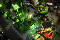 Foto: Eine Nahaufnahme eines optischen Labors mit mehreren Geräten und grünen Laserstrahlen. Verschiedene Linsen und Spiegel sind auf einer perforierten Metallplatte montiert. Ein Gerät zeigt eine Skala mit der Aufschrift "B. Halle". Kabel und Schläuche sind ebenfalls sichtbar.