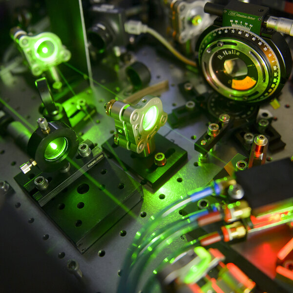 Foto: Eine Nahaufnahme eines optischen Labors mit mehreren Geräten und grünen Laserstrahlen. Verschiedene Linsen und Spiegel sind auf einer perforierten Metallplatte montiert. Ein Gerät zeigt eine Skala mit der Aufschrift "B. Halle". Kabel und Schläuche sind ebenfalls sichtbar.