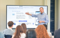 Eine Person steht vor einer Gruppe und präsentiert Informationen auf einem großen Bildschirm. Der Text auf dem Bildschirm beschreibt ein Forschungsprojekt mit Details zur Teilnahme. Im Hintergrund sind mehrere sitzende Personen zu sehen, die der Präsentation folgen.