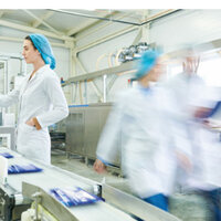 Foto: Eine Person in weißem Kittel und blauer Haube bedient ein Kontrollpanel in einer Fabrikumgebung. Im Vordergrund laufen zwei weitere Personen in ähnlicher Kleidung an einer Produktionslinie vorbei. Maschinen und Förderbänder sind sichtbar.