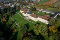 Foto: Luftaufnahme von Schloss Hohenheim, einem großen historischen Gebäude mit symmetrischem Design und Kuppeldach. Es ist von gepflegten Rasenflächen und Bäumen umgeben. Im Hintergrund sind weitere Gebäude und Felder zu sehen. Wege führen durch die Grünflächen.