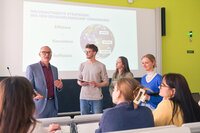 Foto: Eine Gruppe von Personen steht in einem Hörsaal vor einer Präsentation mit dem Titel 'Nachhaltigkeits-Strategien: Wie den Ressourcenbedarf verringern?'. Auf der Folie sind die Begriffe Effizienz, Konsistenz und Suffizienz zu sehen.
