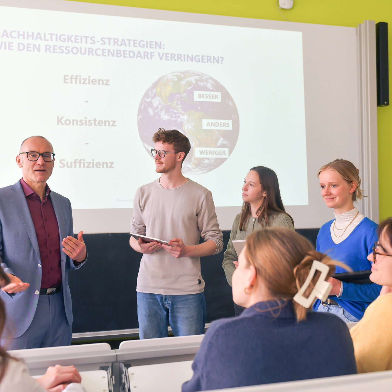 Foto: Eine Gruppe von Personen steht in einem Hörsaal vor einer Präsentation mit dem Titel 'Nachhaltigkeits-Strategien: Wie den Ressourcenbedarf verringern?'. Auf der Folie sind die Begriffe Effizienz, Konsistenz und Suffizienz zu sehen.