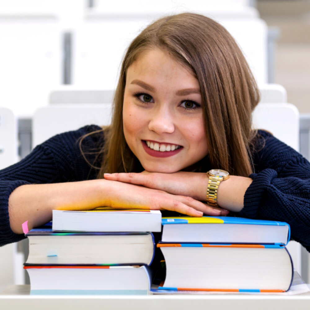 Junge Studentin lehnt lächelnd auf einem Stapel Bücher im Hörsaal.