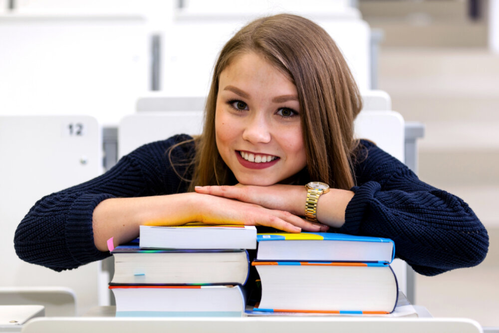 Junge Studentin lehnt lächelnd auf einem Stapel Bücher im Hörsaal.