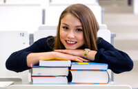 Junge Studentin lehnt lächelnd auf einem Stapel Bücher im Hörsaal.
