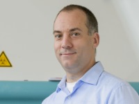 Foto: Eine Person in einem hellblauen Hemd steht vor einer Maschine. Im Hintergrund ist ein gelbes Warnschild mit einem schwarzen Ausrufezeichen sichtbar. Die Umgebung wirkt industriell oder technisch.